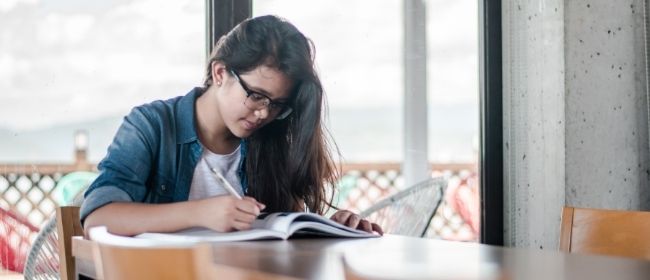 Teenage girl sitting at a table in front of a large window while writing in her notebook.