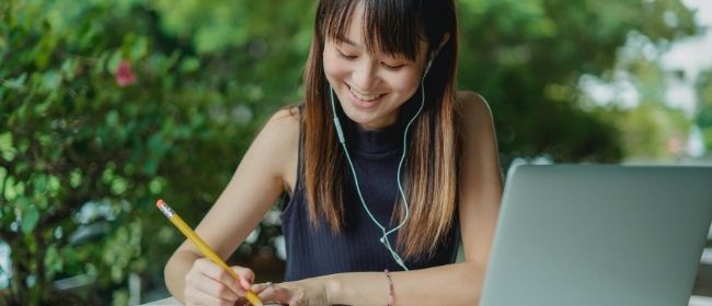 Young girl studying on her computer while writing in a notebook.