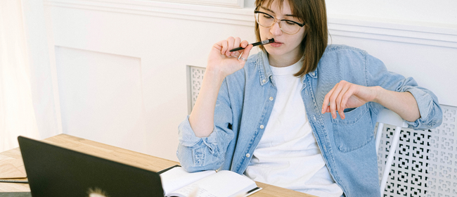 Contemplative young woman biting on pen while sitting in front of an open book and laptop.