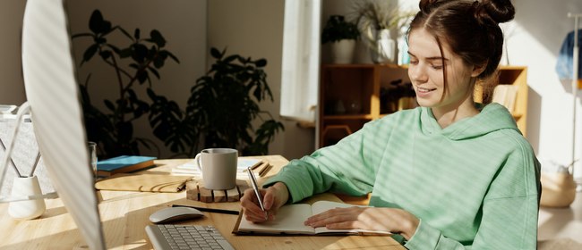 Teenage girl sitting in front of a computer at a desk, while writing in a notebook.