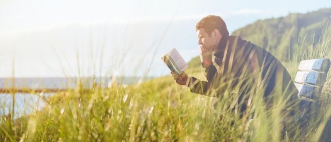 Man reading book on bench outside in grass.