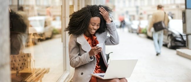 A young woman sitting on a bench while looking at her phone and laptop