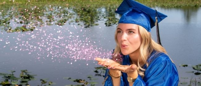 Girl in graduation cap and gown blowing glitter in front of a lake.