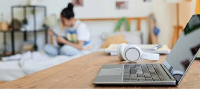 Teen playing on ukelele in background with laptop and desk in the foreground.