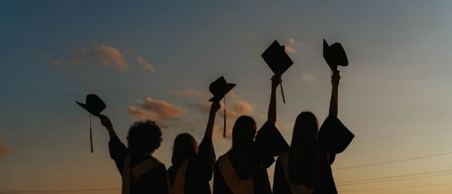 Students with their backs facing the camera wearing graduation robes and holding graduation caps in the air.