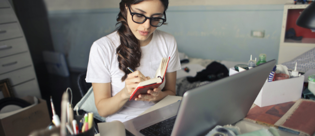 Girl with glasses writing in notebook, sitting at a cluttered desk with laptop open.