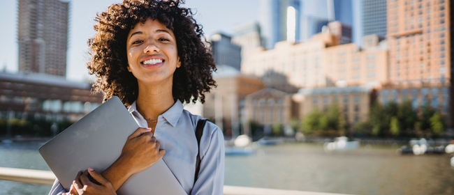 Young woman smiling and looking proud while holding laptop in front of a blurred city background.