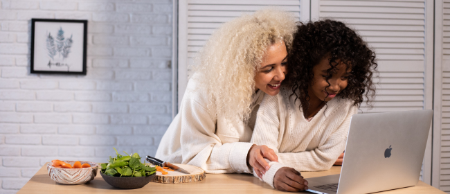 Excited mother and her daughter lean on a table full of snacks and look at a laptop screen.